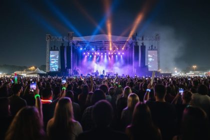 Nighttime view of a large outdoor music festival crowd facing a brightly lit main stage with colorful spotlights and light haze.